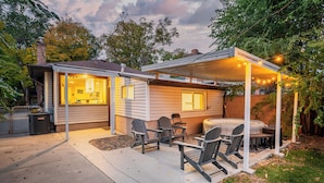 A covered outdoor patio with chairs and a table, a small house with a visible kitchen, and a string of lights. - Relaxing & Stylish: Close to Downtown ~ Yard ~ Pkg (Salt Lake City)