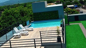Un área de piscina en la azotea con sillones de descanso, un cielo azul despejado y vistas a montañas distantes.