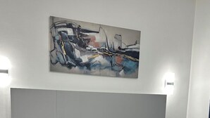 Un dormitorio moderno con una cama grande, ropa de cama blanca, un plaid negro y un cuadro sobre la cama. Hay dos mesitas de noche y lámparas de pared.