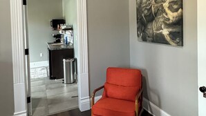 Un couloir avec un plancher en bois, une chaise rouge, un tableau accroché au mur et une porte donnant sur un espace cuisine.