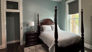 Une chambre à coucher avec un lit à baldaquin, une table de chevet avec une lampe, un ventilateur de plafond et une grande fenêtre donnant sur l’extérieur.