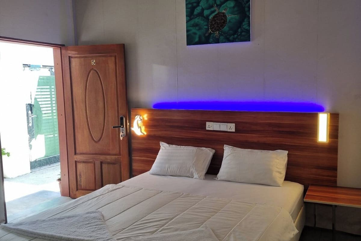 Een slaapkamer met een houten hoofdbord, twee witte kussens, een bed met een witte deken, een nachtkastje met een lamp en een deur met een nummer.