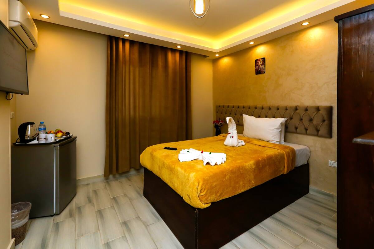 A hotel room with a bed, a small desk with a kettle, a trash bin, and a television mounted on the wall.