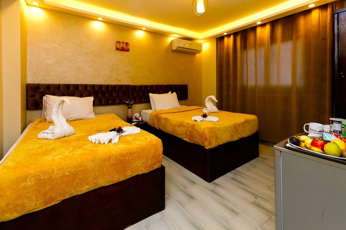 A hotel room with two beds, each with a golden blanket and a swan-shaped pillow. There is a small table with a fruit bowl and a mug.