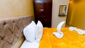 Una habitación de hotel con un cabecero capitonado, una cama con almohadas blancas, una colcha naranja y una toalla doblada sobre la cama.