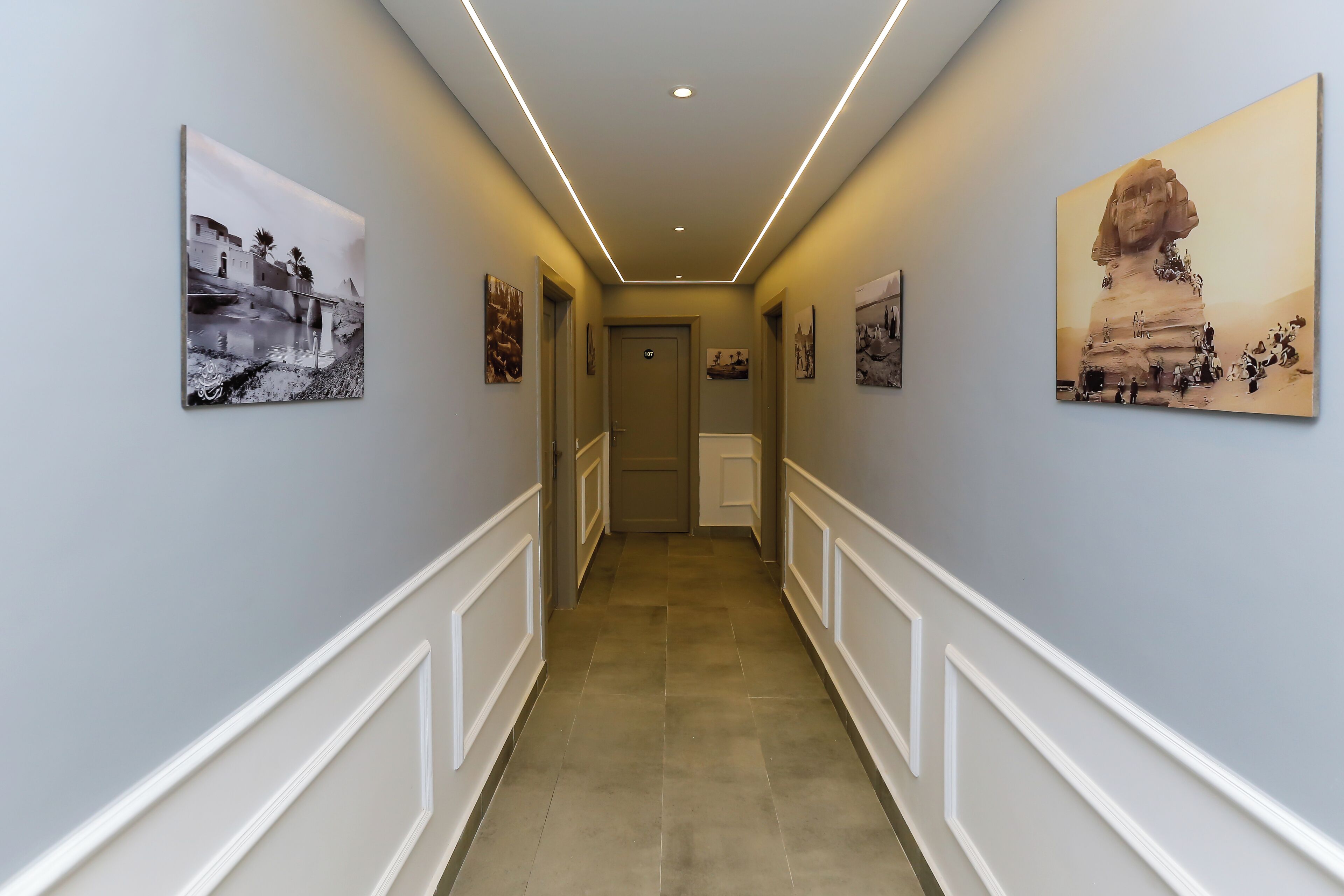 A hallway with a light blue wall, white trim, and a tiled floor. There are framed pictures on the walls and a door at the end.