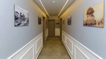 A hallway with a light blue wall, white trim, and a tiled floor. There are framed pictures on the walls and a door at the end.