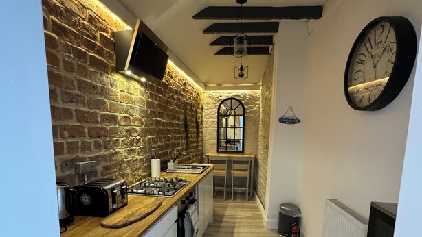 A narrow kitchen with a brick wall, wooden counter, and a clock on the wall. - Angel Mews - Westend Glasgow Uni Cottage (Glasgow)