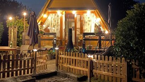 A wooden cabin with a triangular roof, a porch with chairs and a table, and a wooden fence surrounding the property. - Renoviertes Nurdachhaus in Ostfriesischer Natur am Wasser für Familien (Südbrookmerland)