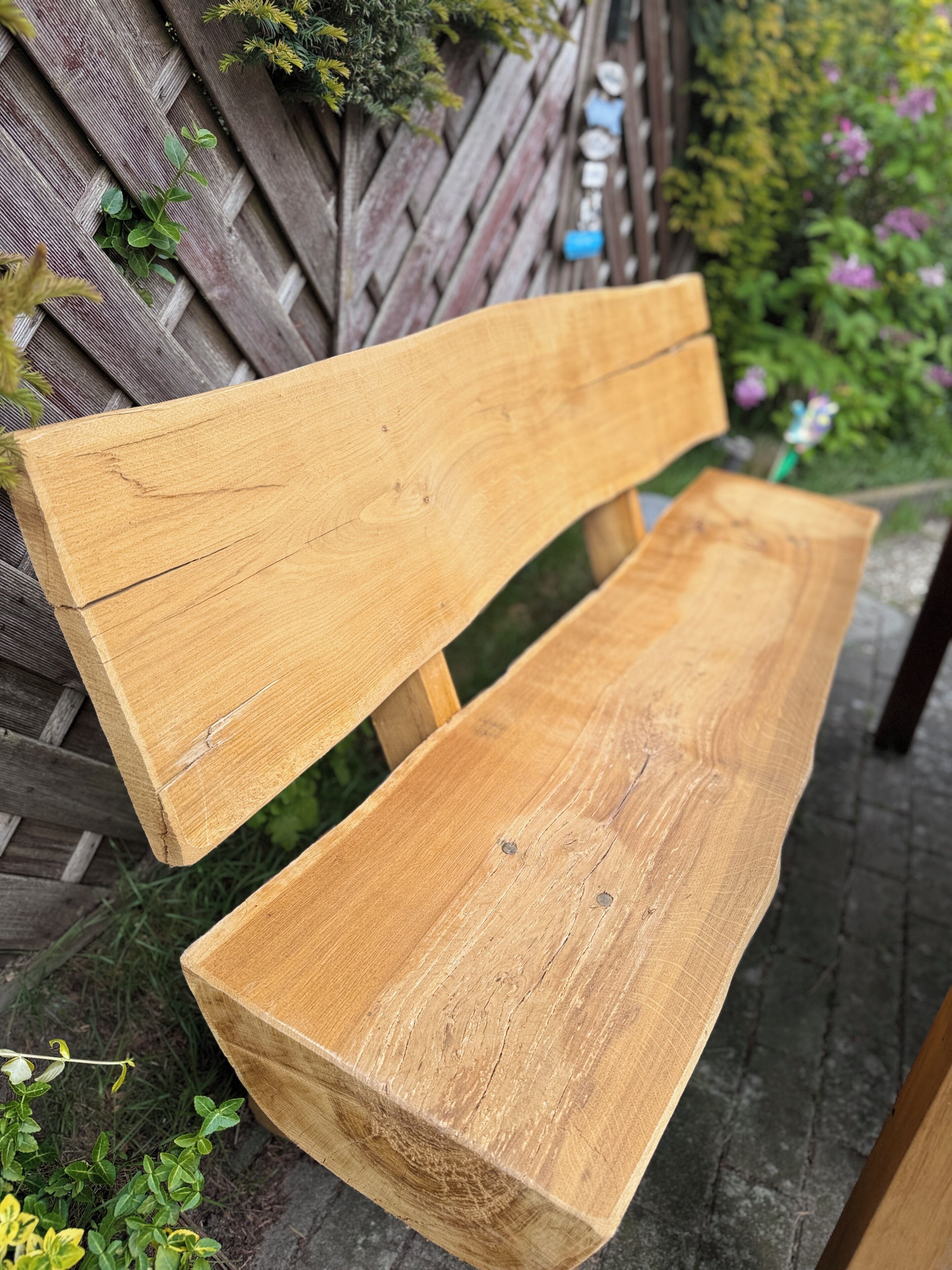 Un banco de madera con respaldo de listones y asiento plano, ubicado al aire libre frente a una valla de madera y follaje en el fondo.
