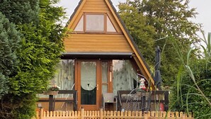 A wooden house with a triangular roof, a wooden fence, and a green boat docked in front. - Renoviertes Nurdachhaus in Ostfriesischer Natur am Wasser für Familien (Südbrookmerland)