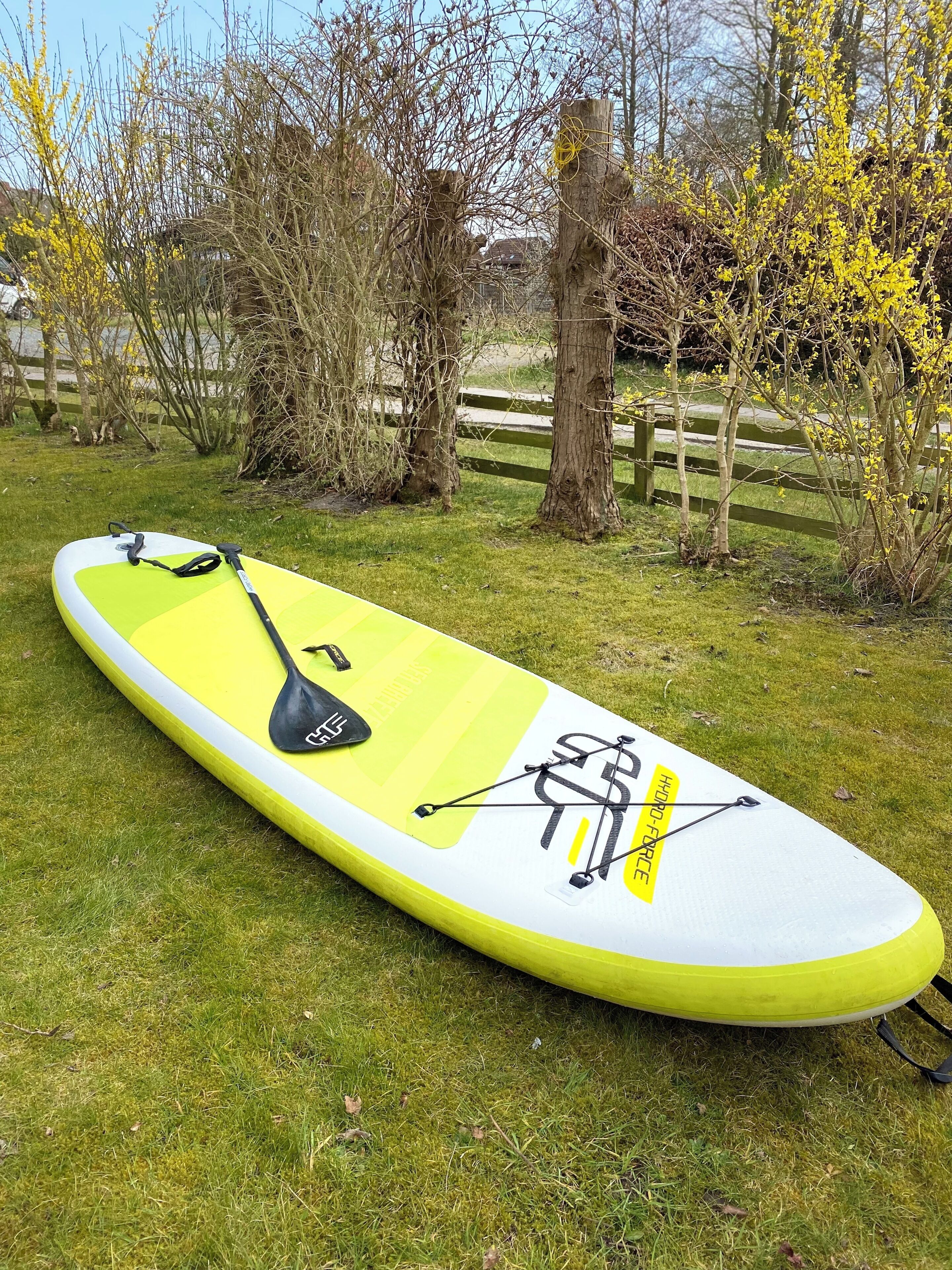 Una tabla de paddleboard con diseño amarillo y blanco, una remo y una cuerda sobre una zona cubierta de césped.
