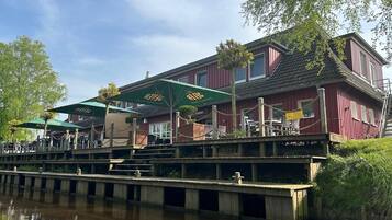 Een restaurant aan de rivier met terras, een houten dek en een rood gebouw.