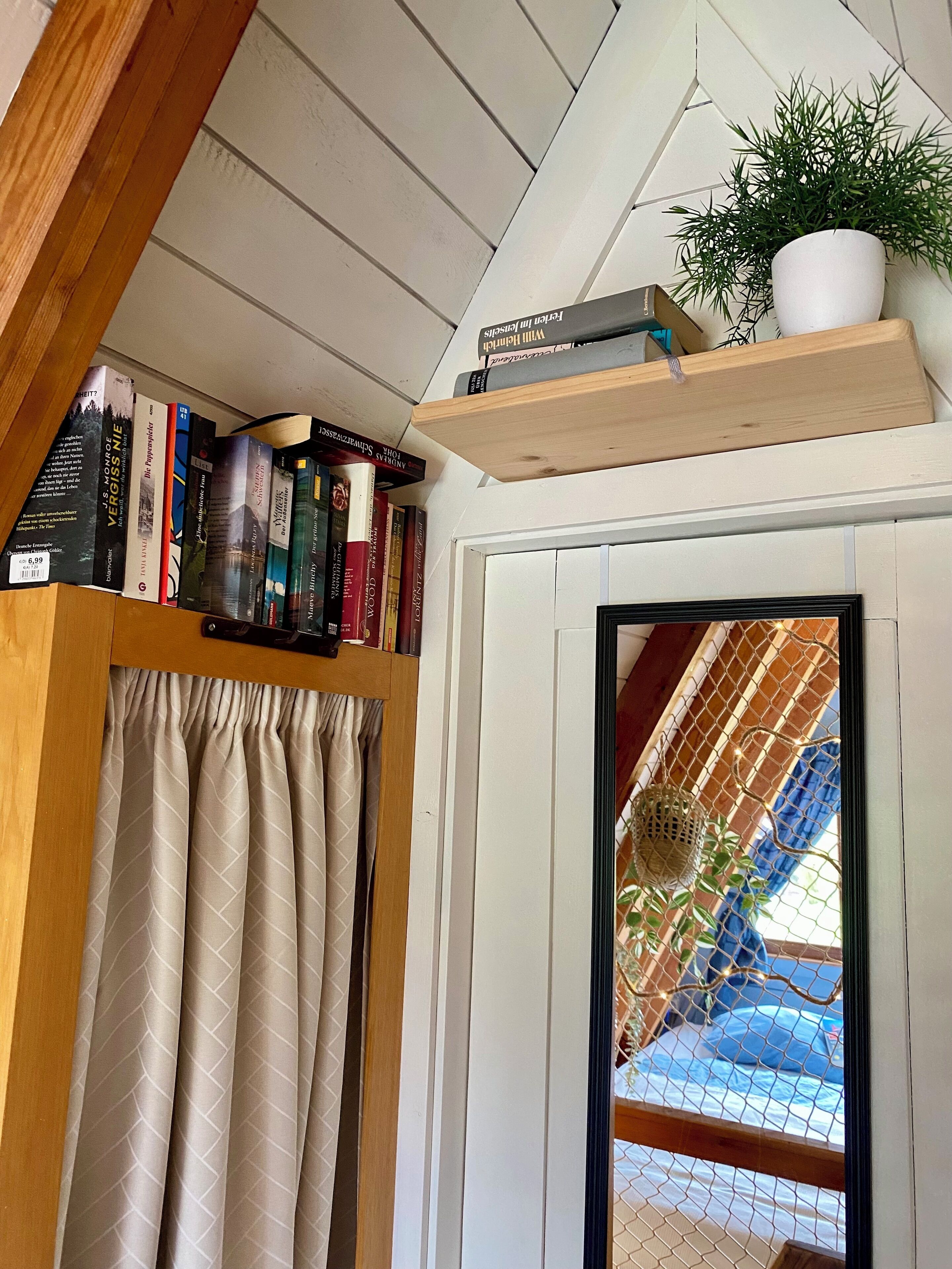 A room with a wooden ceiling, a bookshelf with books, a potted plant, and a view through a window with a curtain.
