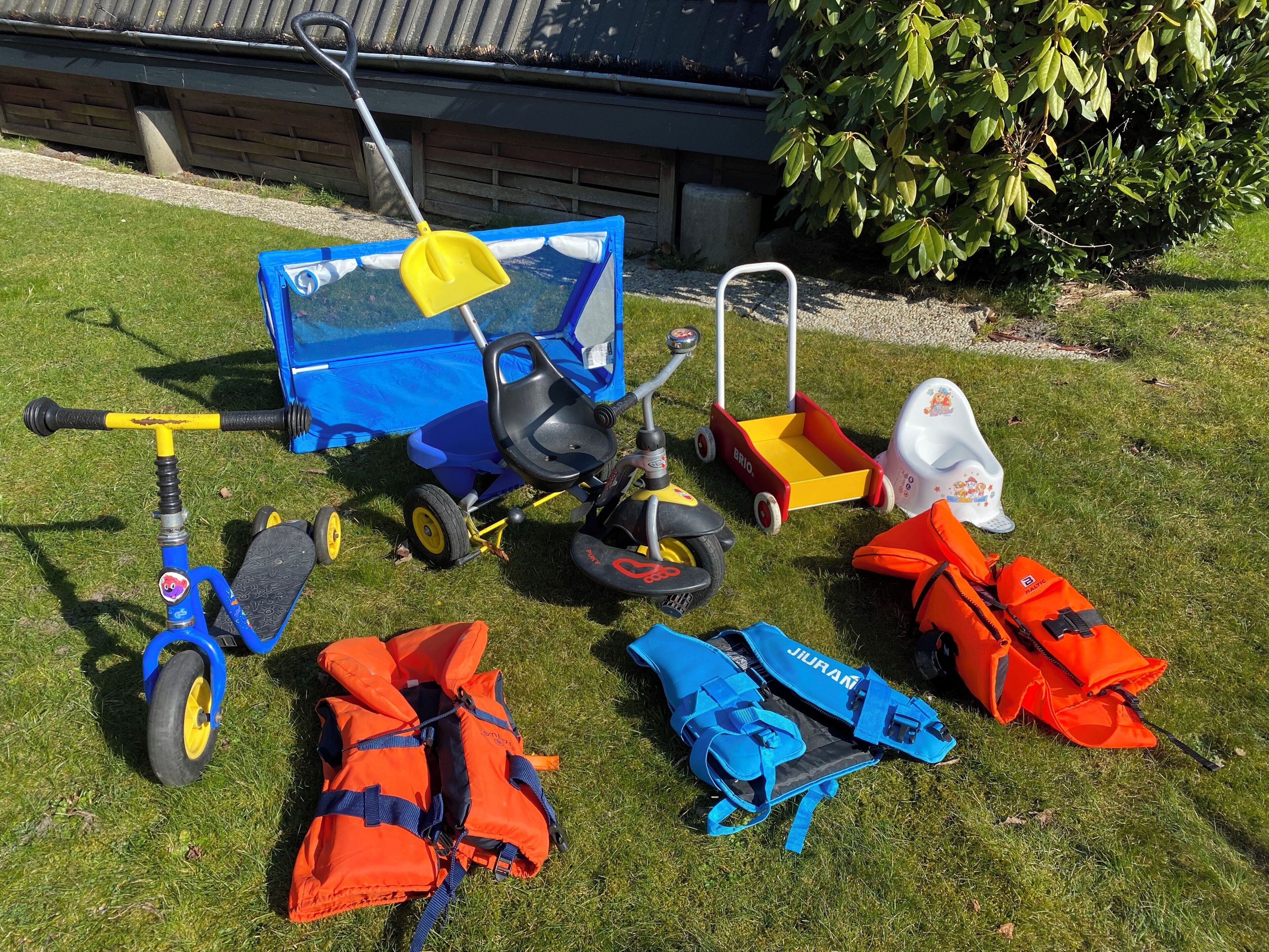 Een gazon met diverse kinderspeelgoed en veiligheidsuitrusting, waaronder een driewieler, een autospeeltje en reddingsvesten.