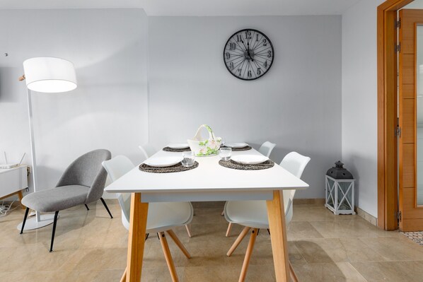 A dining area with a white table set for four, surrounded by white chairs. A modern lamp and a wall clock are visible.
