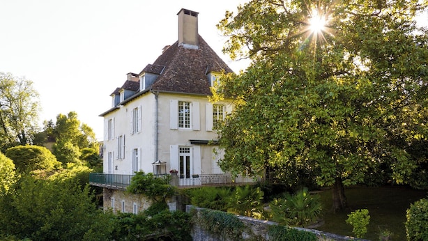 A large, white building with a steep roof, surrounded by lush greenery and a stone wall.