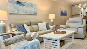 A living room with a beige sofa, a white armchair, a coffee table, and a rug. There are two lamps on side tables, and artwork on the walls. - Beachfront Sanibel Escape – Steps to Sand & Sunset Views (Sanibel)