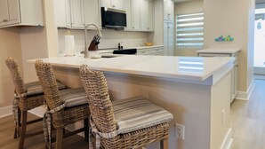 A modern kitchen with white cabinetry, a central island with bar stools, and a large window with blinds. - Beachfront Sanibel Escape – Steps to Sand & Sunset Views (Sanibel)
