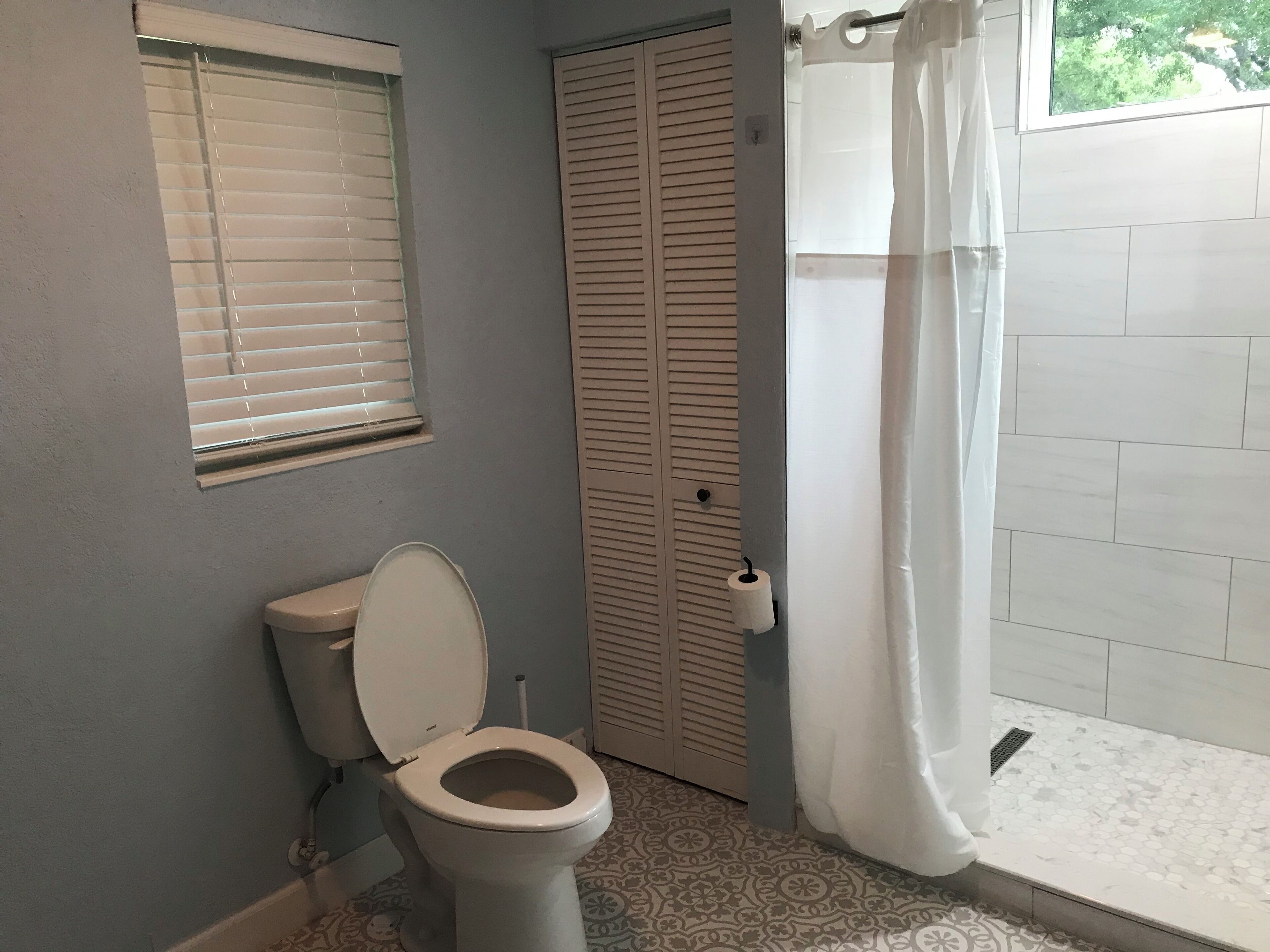 A bathroom with a toilet, a window with blinds, a shower with a curtain, and a patterned floor.