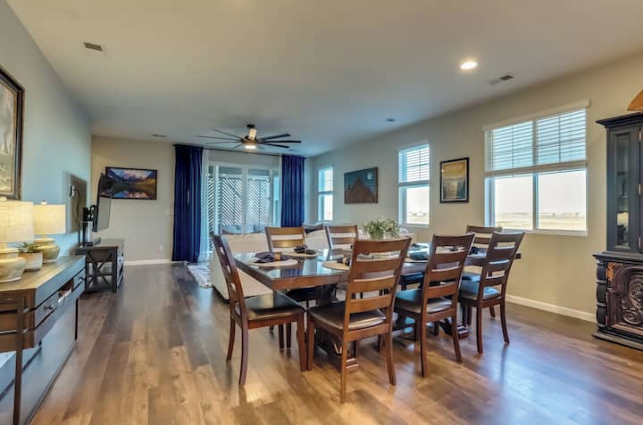 A spacious dining area with a wooden table and chairs, a ceiling fan, and large windows with blinds.