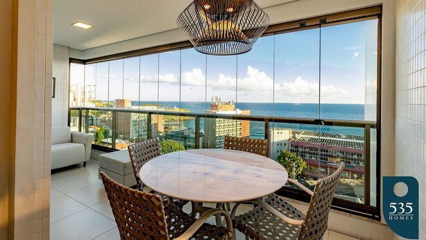 Ein modernes Wohnzimmer mit rundem Tisch und Korbstühlen, großen Fenstern mit Blick auf Stadt und Meer, sowie einer dekorativen Pendelleuchte.