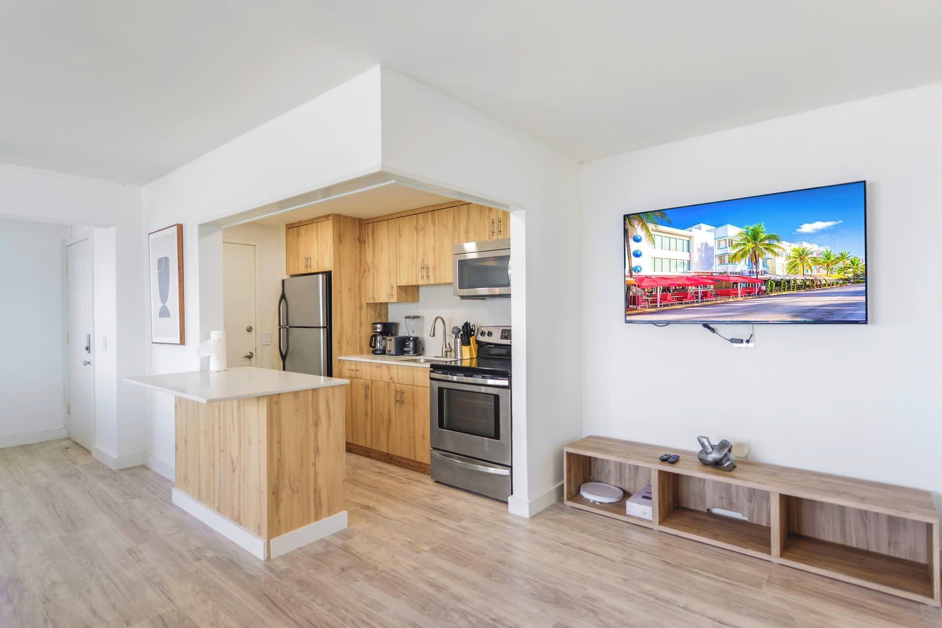 Eine moderne Küche mit Holzschränken, Edelstahlgeräten und einem eingebauten Backofen. Eine kleine Holzablage mit einem Fernseher, der eine Strandansicht zeigt.