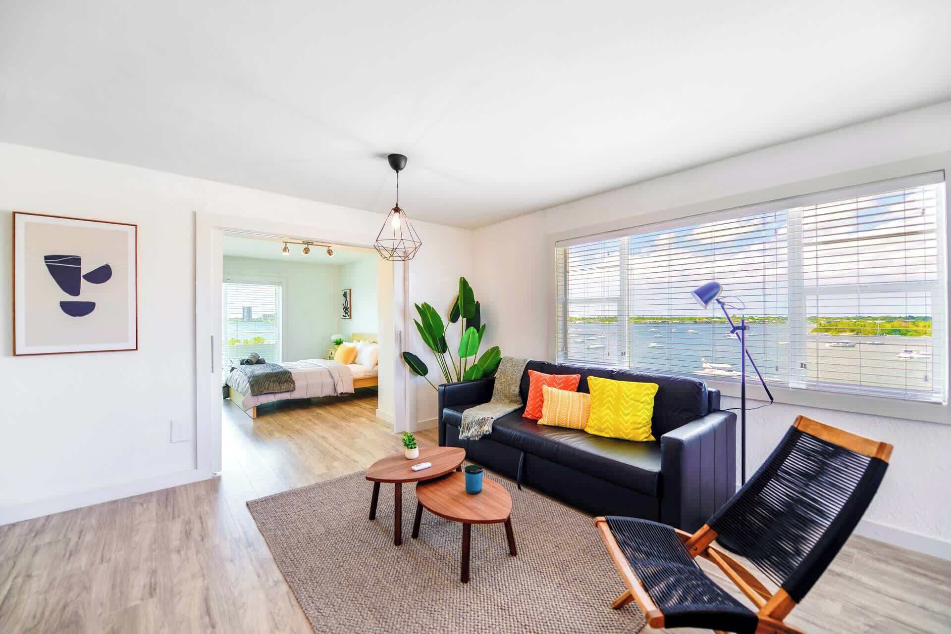 A modern living room with a sofa, a coffee table, and a chair. There is a large window with blinds, a ceiling light, and a framed picture on the wall.
