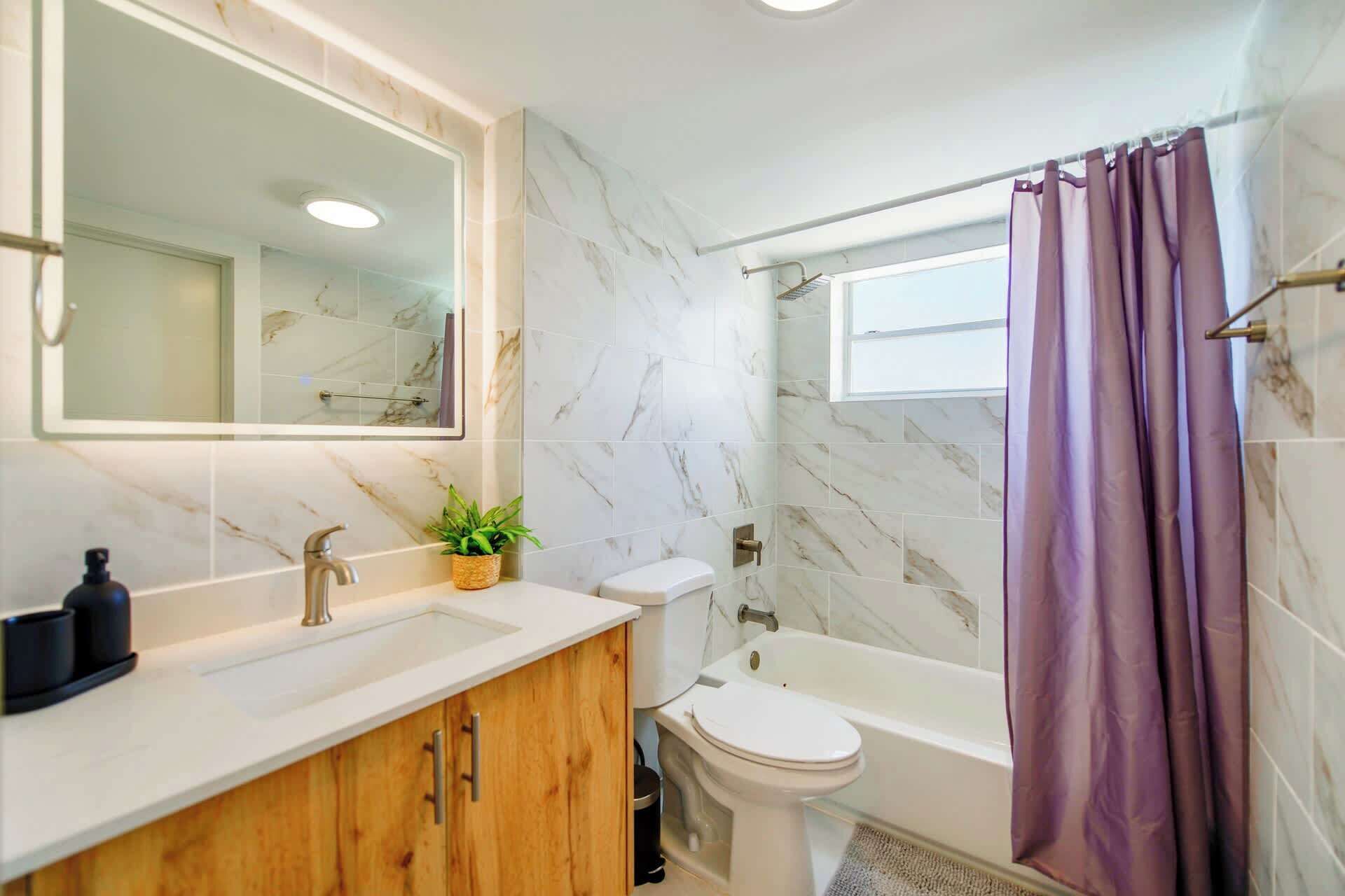 A bathroom with a marble-tiled wall, a white sink, a wooden vanity, a toilet, and a bathtub with a purple shower curtain.