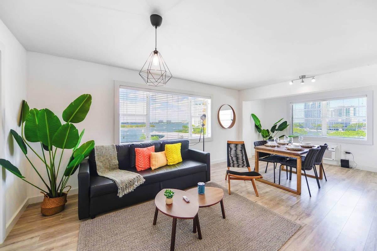 Uma sala de estar moderna com um sofá preto, almofadas coloridas, uma mesa de centro redonda e uma área de jantar com mesa e cadeiras de madeira.