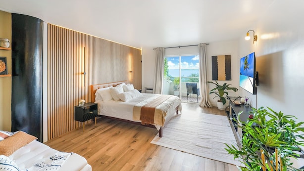 A modern bedroom with a large bed, a bedside table, a desk with a chair, a potted plant, and a view of the outdoors. - Stunning Studio Bay Views & Parking (North Bay Village)