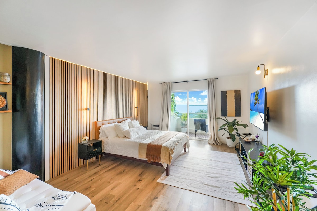 Quarto moderno com uma cama grande, mesa de cabeceira, escrivaninha com cadeira, planta em vaso e vista para o exterior.