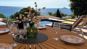 A wooden table set for dining with a view of the sea, a vase with dried flowers, and a bottle of water.
