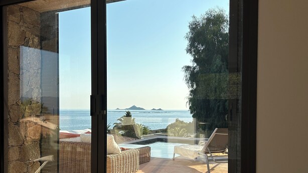 A modern room with a large sliding glass door leading to an outdoor area with a pool, lounge chairs, and a view of the sea.