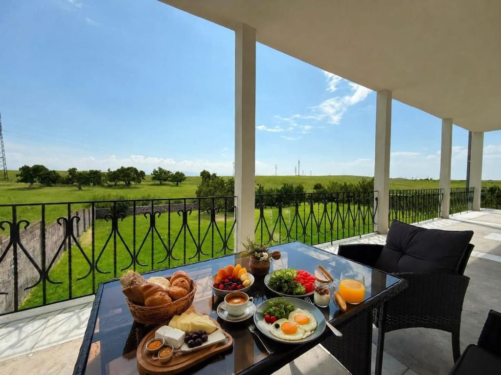 Une table de petit-déjeuner dressée sur un balcon, offrant une vue sur un champ verdoyant et un ciel dégagé, et sur laquelle on trouve une variété d’aliments ainsi qu’un verre de jus d’orange.