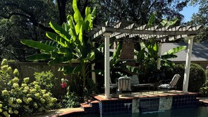 Een helder zwembad met een bakstenen rand, omringd door weelderige begroeiing en een houten pergola.