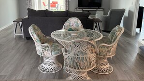 A modern living room with a grey sofa, a TV, and a dining area with wicker chairs and a glass table. - Charming 2-bedroom house in fabulous Hawkins Village of The Villages Florida. (Wildwood)