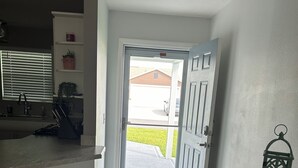 A hallway with a door leading outside, a kitchen area with a sink and countertop, and a small shelf with a plant. - Charming 2-bedroom house in fabulous Hawkins Village of The Villages Florida. (Wildwood)