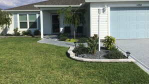 A two-story house with a light blue garage door, a front porch with a bench, and a well-maintained lawn. - Charming 2-bedroom house in fabulous Hawkins Village of The Villages Florida. (Wildwood)