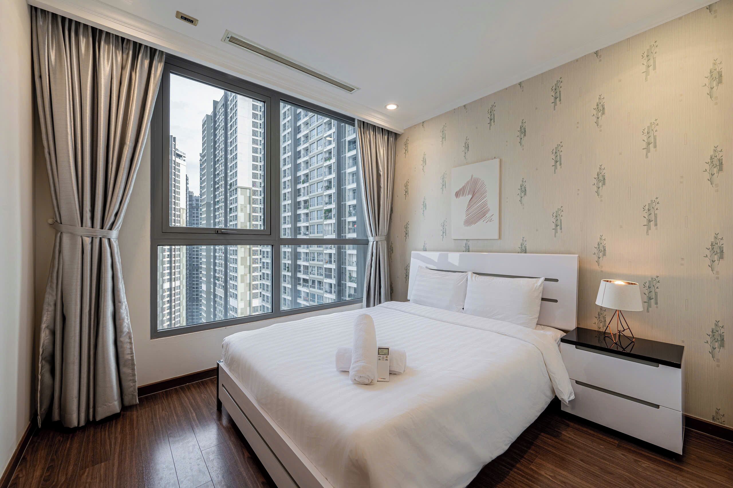 A hotel room with a large window, a neatly made bed, a bedside table with a lamp, and a wall with floral wallpaper.