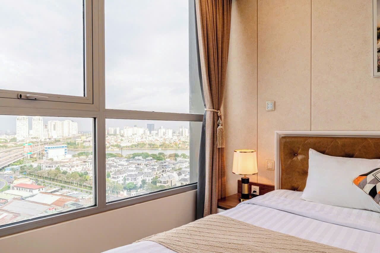 Habitación de hotel con un ventanal grande, cama con sábanas blancas, mesita de noche con lámpara y vistas a un paisaje urbano.