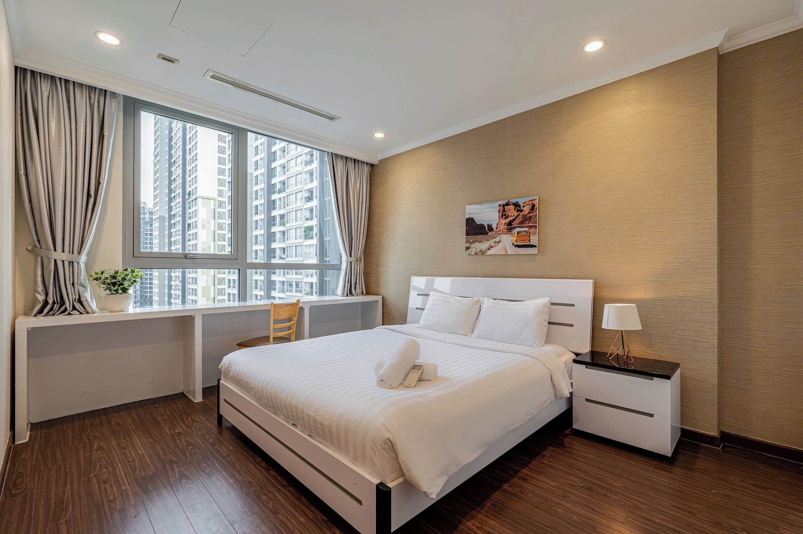 A hotel room with a large bed, a bedside table with a lamp, a window with curtains, and a view of tall buildings.