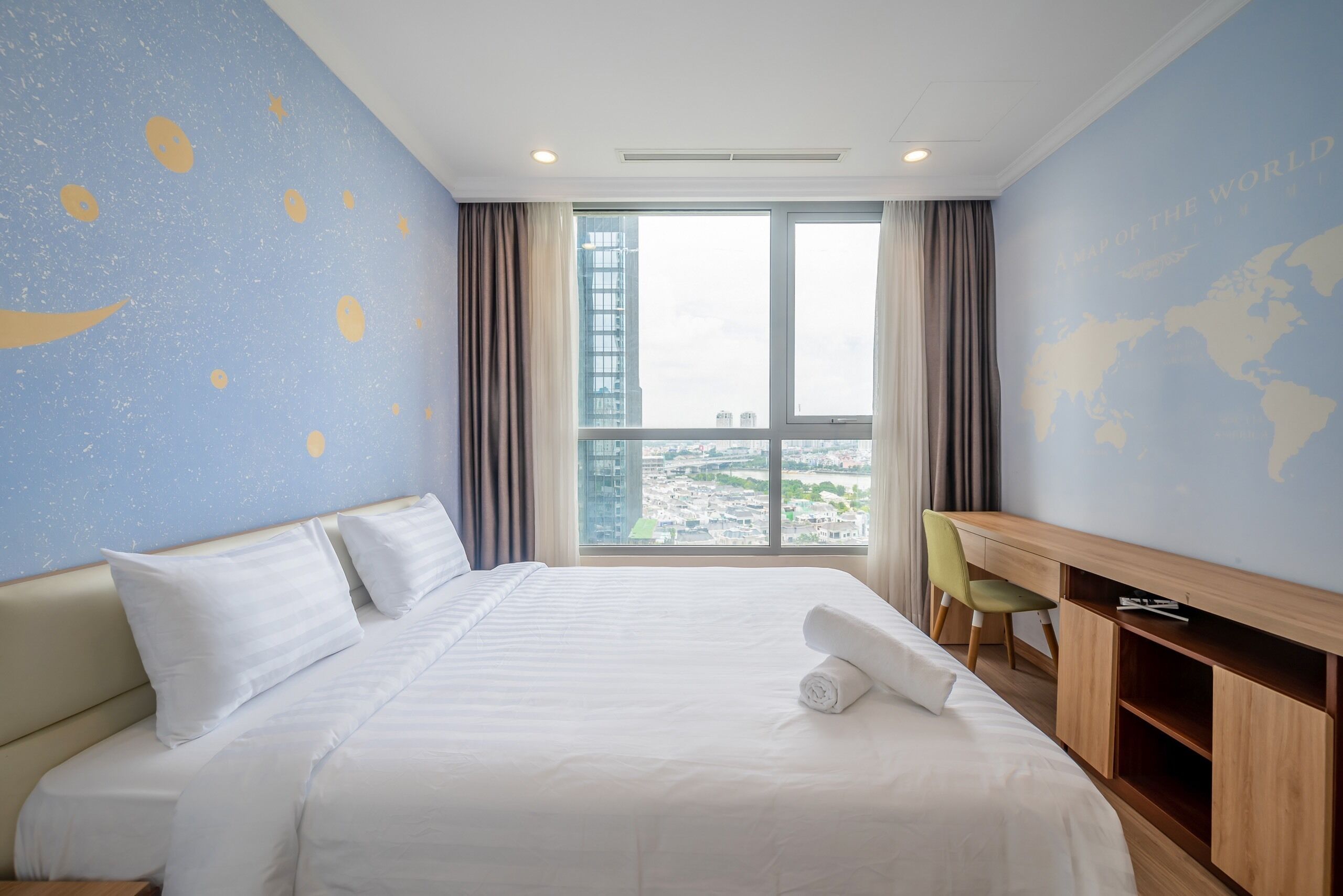 A hotel room with a large bed, a desk, and a view of a cityscape through a large window.