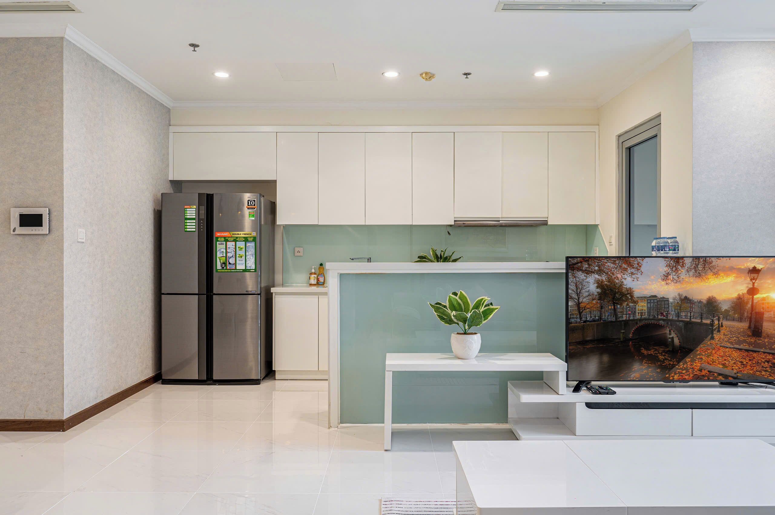 A modern kitchen with white cabinets, a stainless steel refrigerator, a small island with a plant, and a TV displaying a scenic view.