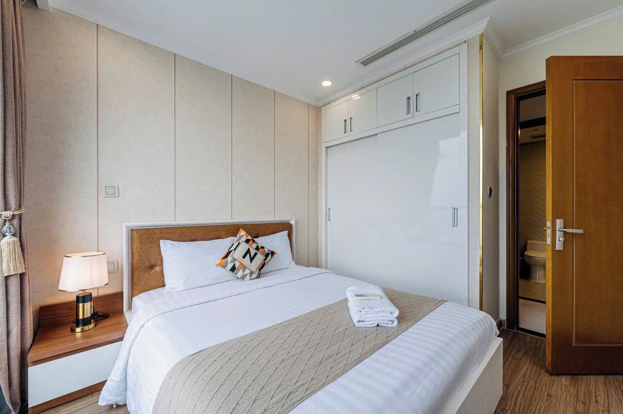 A hotel room with a bed, bedside table, lamp, wardrobe, and a door leading to a bathroom.