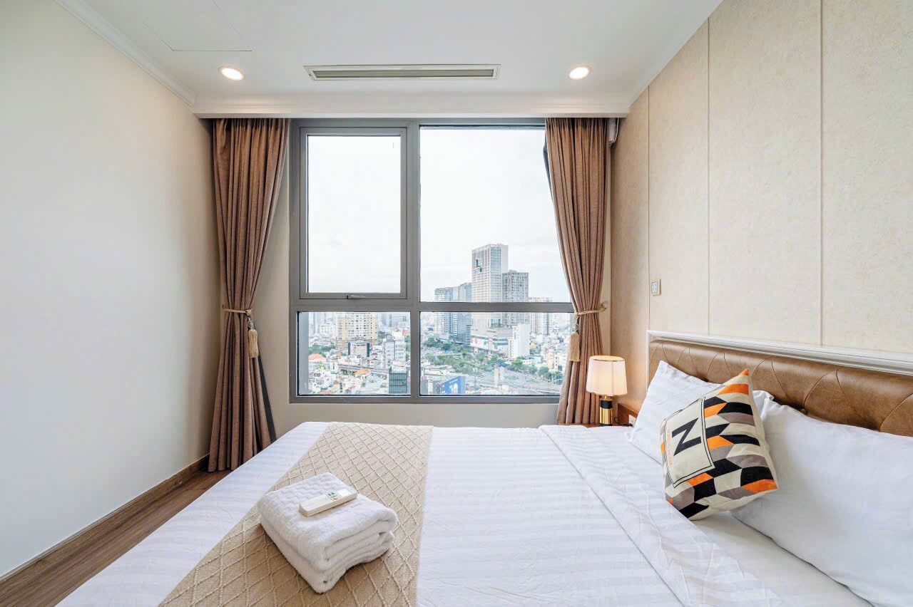 A hotel room with a large window, a bed with white linens, a headboard, and a bedside table with a lamp.