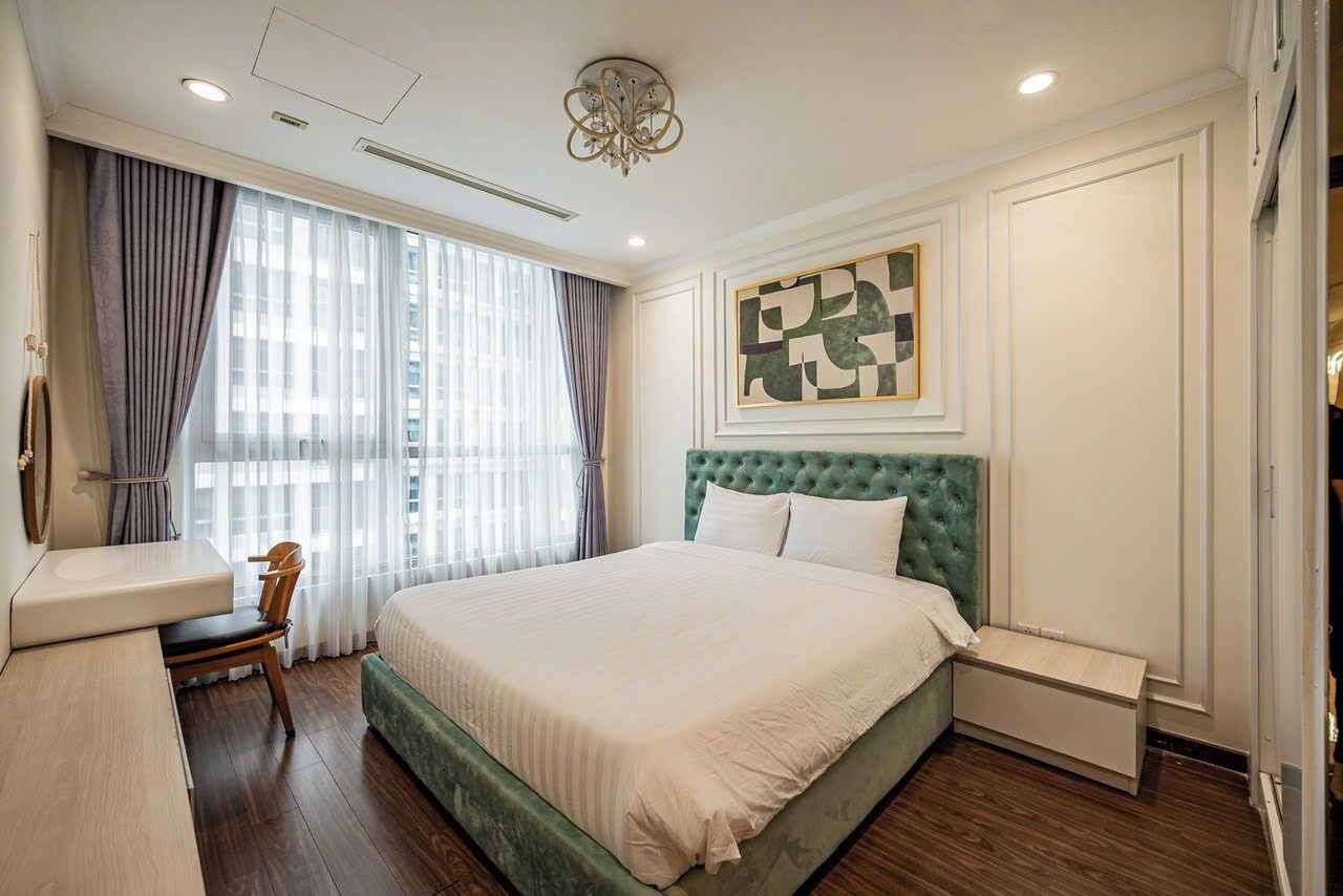 A hotel room with a green upholstered bed, a bedside table, a chair, and a large window with curtains.