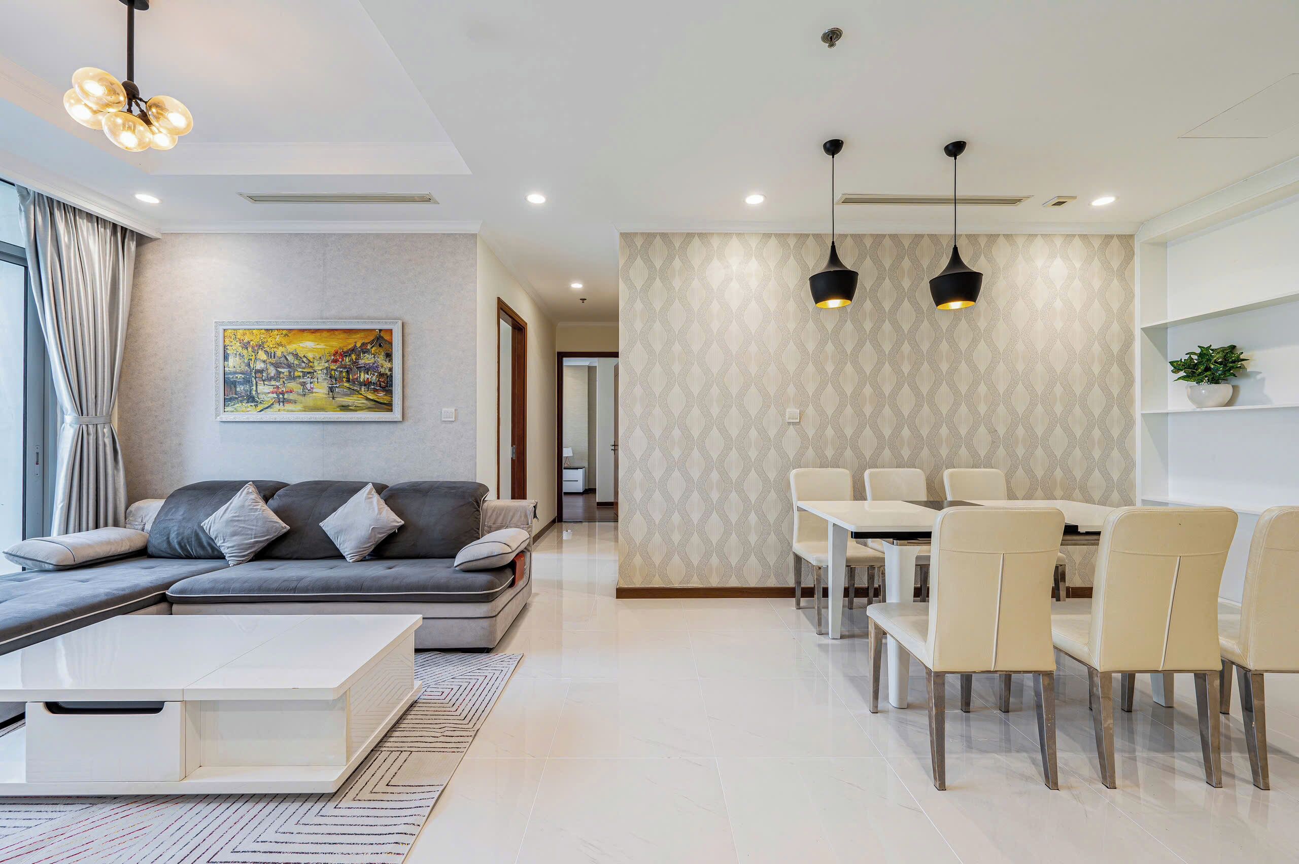 A modern living room with a grey sofa, a white coffee table, and a dining area with beige chairs and a table.