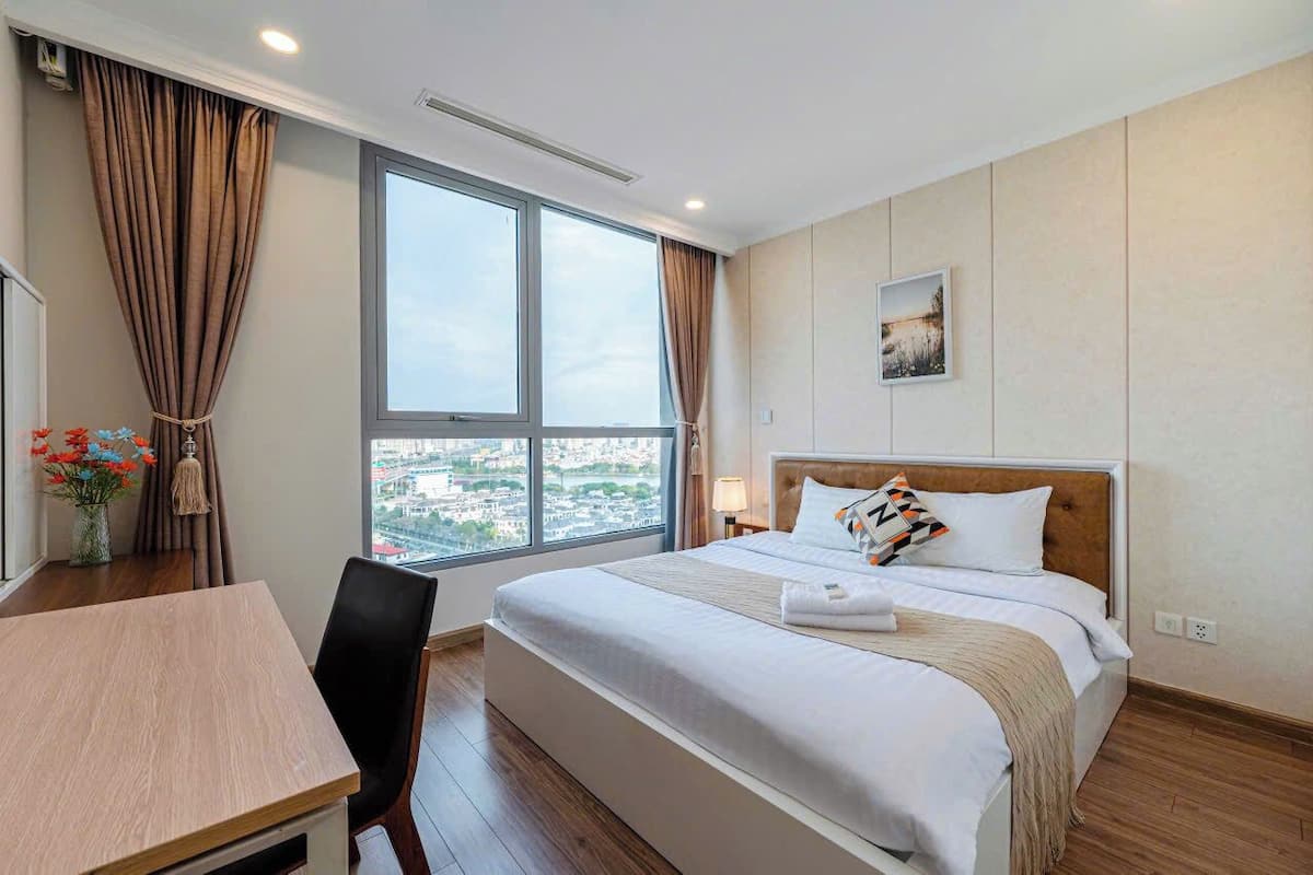 Quarto de hotel com uma janela grande, uma cama com roupa branca, uma mesinha de cabeceira e uma cadeira.