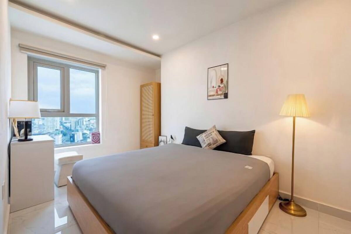Un dormitorio moderno con una cama grande, una mesita de noche, una lámpara y una ventana con vistas a la ciudad.